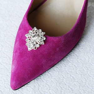 May include: Close-up of a fuchsia suede pointed-toe shoe. A silver-colored, ornate floral embellishment with clear crystals adorns the vamp. The shoe's design is elegant and suitable for formal occasions.