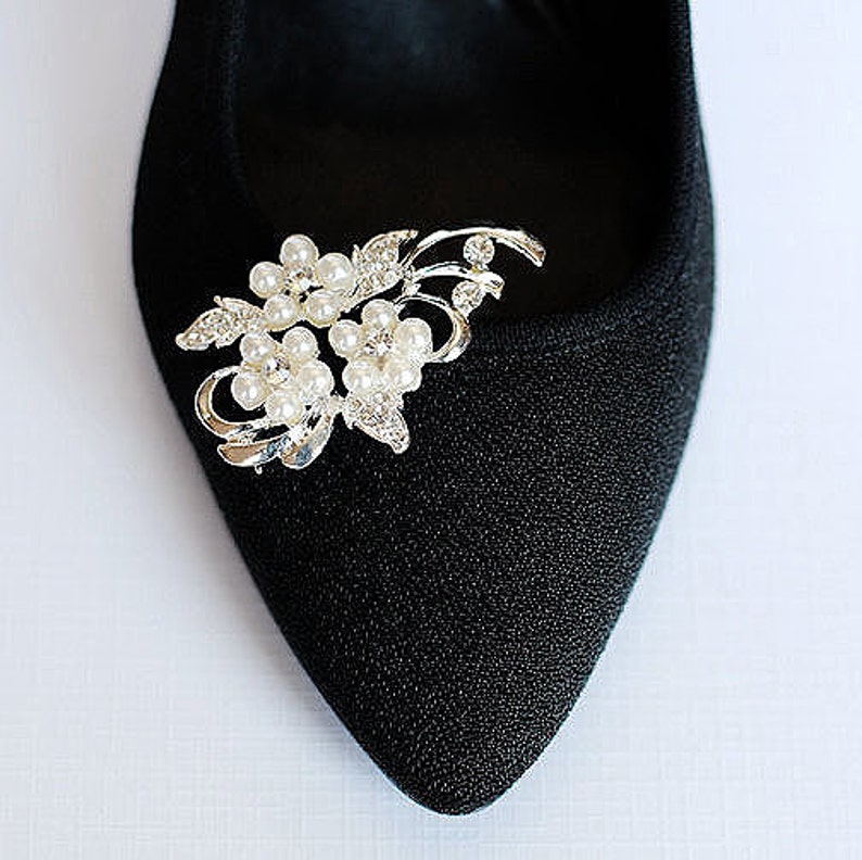 May include: A silver shoe clip with a floral design featuring faux pearls and rhinestones. The clip is attached to a black shoe.
