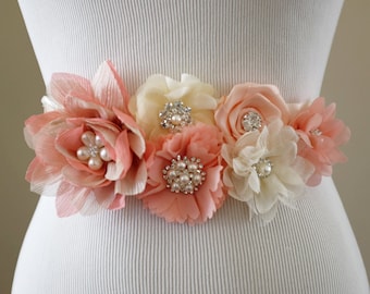 May include: A peach, cream and white floral sash with pearl and rhinestone embellishments. The sash is made of fabric flowers and is tied around a white dress form.
