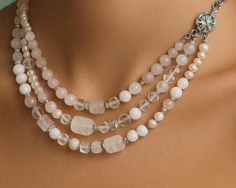 Puede incluir: Collar de tres hilos con cuentas de cuarzo rosa, cristal transparente y perlas blancas. El collar tiene un cierre plateado con un adorno decorativo verde y plateado. Las cuentas son de varias formas y tamaños.