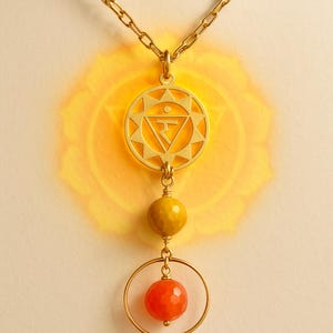 Op de afbeelding: Goudkleurige ketting met een hanger met een zonmotief en een centrale driehoek. De ketting bevat een gele kraal en een oranje, facetgeslepen kraal, die aan een gouden ketting hangen. De achtergrond heeft een zachte gele gloed.