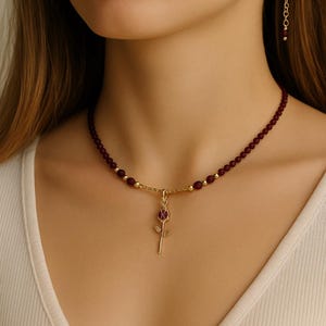 May include: A gold necklace featuring a burgundy beaded chain and a rose pendant. The rose pendant has a burgundy centre and gold petals. A matching burgundy beaded earring hangs from the ear.