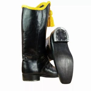 Può includere: Stivali da equitazione in pelle nera con nappa e finiture gialle. Gli stivali hanno un gambale alto e una suola robusta. Il tacco è rinforzato con metallo. Gli stivali sono probabilmente per uso equestre o rievocazioni storiche.