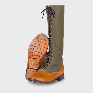 Könnte beinhalten: Ein Paar hohe, olivgrüne und hellbraune Stiefel mit Schnürdesign. Die Stiefel haben eine Ledersohle mit Metallstollen und ein Obermaterial aus Segeltuch. Die Stiefel sind für Outdoor-Aktivitäten konzipiert.