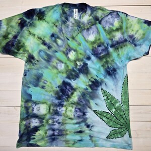 Może przedstawiać: Koszulka typu tie-dye w odcieniach niebieskiego, zielonego i fioletowego. Duży zielony liść konopi indyjskich znajduje się po prawej stronie u dołu. Koszulka ma okrągły dekolt i krótkie rękawy.