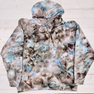 May include: A long-sleeve hooded sweatshirt featuring a tie-dye pattern in shades of brown, tan, black, and light blue. The garment includes a front kangaroo pocket, a hood with matching drawstrings, and ribbed cuffs at the sleeves and hem.