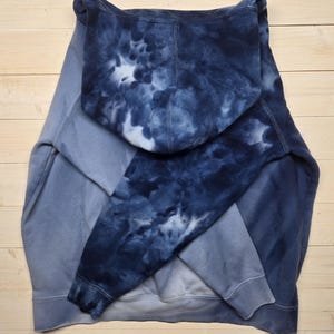 May include: A blue tie-dye hoodie with a hood and long sleeves. The hoodie is a mix of light and dark blue shades, with a prominent tie-dye pattern on the hood and sleeves. The garment is made of a soft material.