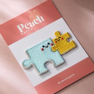 May include: Two embroidered puzzle piece patches, one light blue and one yellow, with smiling faces and gold trim. The patches are on a pink and white card with "Peach Patch" text and the Instagram handle @_peachpatch.