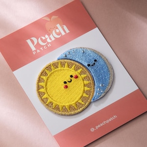 Op de afbeelding: Geborduurde patches met een lachende gele zon en een lachende blauwe maan. De patches liggen op een roze en witte kaart met de tekst "Peach Patch". De zon heeft een gouden rand en rode wangen. De maan heeft een zilveren rand.