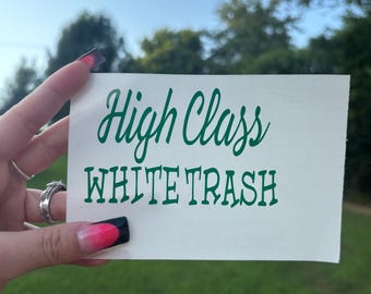 May include: A rectangular white vinyl decal featuring the dark green text High Class in a cursive script font positioned above the text WHITE TRASH in a bold, blocky, uppercase sans-serif font. The decal is presented on a white backing sheet.