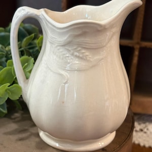 May include: A cream-colored ceramic pitcher featuring a glossy finish with a subtle crackle texture. The pitcher has a curved handle, a scalloped rim, and an embossed wheat stalk design wrapping around the upper body of the vessel.