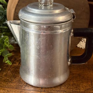 May include: A silver-colored aluminum percolator coffee pot with a black handle and a lid featuring a clear glass knob. The coffee pot has a tapered body, a side spout, and a metallic finish with visible surface wear and light scratches.