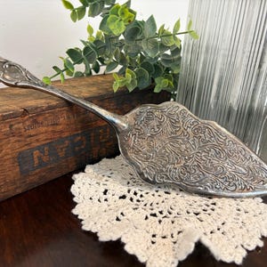 May include: Silver cake server with ornate floral design. The server rests on a cream-colored, crocheted doily. A wooden box and a clear glass vase are in the background. The server's blade is approximately 4 inches wide.