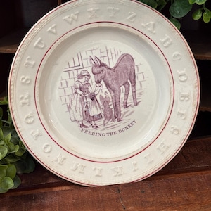 May include: A round ceramic plate with a cream-colored surface and a red rim. The border features embossed letters of the alphabet. The center displays a purple illustration of two children feeding a donkey, with the text FEEDING THE DONKEY.