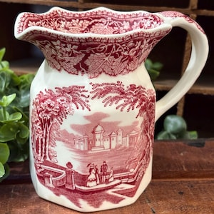 May include: A cream-colored ceramic pitcher featuring a deep red transferware pattern. The design depicts a castle landscape with figures in the foreground, surrounded by a decorative border of leaves and vines along the rim and handle.