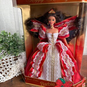 May include: A Barbie doll in a box, wearing a red and white gown with gold accents. The doll has dark hair and is adorned with a tiara. The box reads "Happy Holidays" and "Special Edition Barbie".