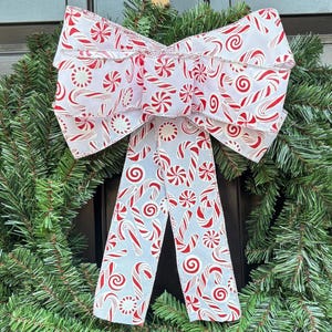 Pode incluir: Uma grinalda de Natal com agulhas de pinheiro verdes e um grande laço. O laço tem um fundo azul claro com padrões de bengalas de doces e redemoinhos vermelhos. A grinalda está pendurada numa porta preta.