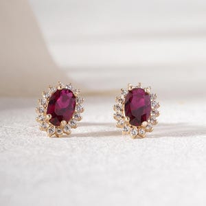 Può includere: Un paio di orecchini a lobo dorati, ciascuno con una grande gemma rossa ovale circondata da gemme chiare più piccole. Gli orecchini sono posti su uno sfondo bianco morbido, evidenziando il design.