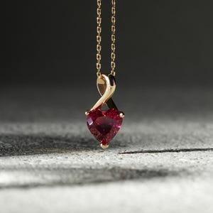 Puede incluir: Un collar dorado con un colgante de piedra preciosa roja oscura en forma de corazón. El colgante cuelga de una delicada cadena. El collar se muestra sobre un fondo gris oscuro.