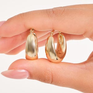 May include: Two gold-colored hoop earrings. The earrings have a rounded, thick design. The earrings are held up by a hand with long, manicured nails. The background is plain white.