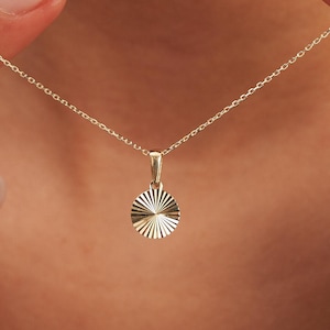 Puede incluir: Un delicado collar de oro con un pequeño colgante redondo con un diseño de rayos de sol. El colgante cuelga de una fina cadena. El collar es de color oro cálido y parece ser de metal.