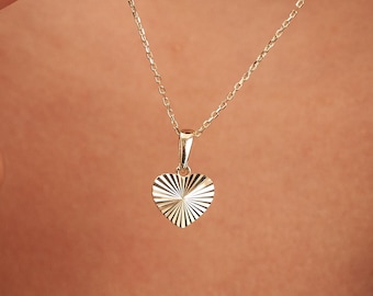 May include: A gold-colored necklace with a heart-shaped pendant. The pendant has a textured, sunburst design. The necklace is delicate and appears to be made of a fine chain. The background is a blurred view of skin.