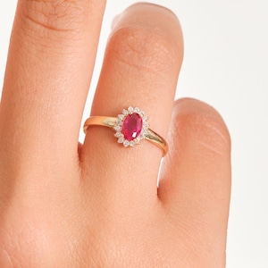 Puede incluir: Un anillo de oro con una gema roja ovalada rodeada de pequeñas gemas blancas. El anillo está en un dedo, con la mano y parte de una cara visibles. La banda del anillo es delgada y dorada.