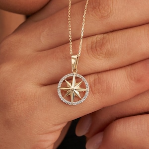 May include: A gold-colored necklace with a compass rose pendant. The pendant features a star design within a circle of small, clear stones. The necklace is held up by a person.
