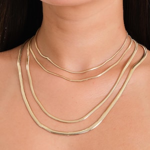 May include: Four gold-coloured chain necklaces of different lengths. The necklaces are layered, featuring a flat, snake-like design. The model is wearing a black vest top. The background is plain.