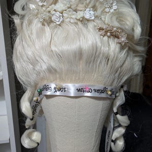 May include: A light blonde wig styled with curls and adorned with white floral accents, pearls, and gold-toned embellishments. The wig is mounted on a mannequin head, with a ribbon around the base that reads "100% Egyptian Cotton".