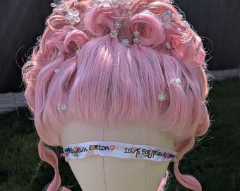 May include: A light pink wig with curled sections and decorative clear flower and gem embellishments. The wig is displayed on a mannequin head, with a ribbon that reads "Egyptian cotton 100% Egyptian".