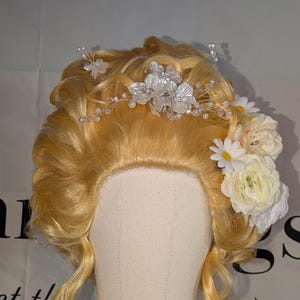 Könnte beinhalten: Blonde Perücke im klassischen Hochsteckstil, verziert mit einem zarten Blumenkranz und weißen Blütengruppen. Die Perücke hat weiche Locken und wird auf einem Schaufensterkopf präsentiert, ideal für Kostüme oder Theater.
