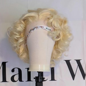 May include: A blonde wig with large curls is displayed on a mannequin head. The wig has a lace front and a band that reads "100% Egyptian cotton". The wig is styled with soft, bouncy curls.