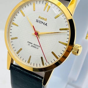 Könnte beinhalten: Eine goldfarbene Armbanduhr mit weißem Zifferblatt und schwarzem Lederarmband. Das Zifferblatt der Uhr hat goldene Zeiger und Markierungen mit dem Text "SONA" und "PARA SHOCK 17 JEWELS". Die Uhr hat eine goldene Krone und ein schwarzes Lederarmband.