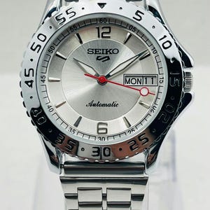 Pode incluir: Relógio automático Seiko prateado com pulseira de aço inoxidável. O mostrador é prateado com detalhes em preto e vermelho, incluindo o dia e a data. A luneta tem marcações pretas de minutos.