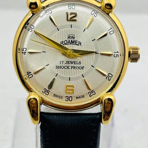Op de afbeelding: Goudkleurig RW Roamer horloge met een zilveren wijzerplaat, zwarte leren band en gouden wijzers. De wijzerplaat toont de tekst "RW ROAMER", "17 JEWELS" en "SHOCK PROOF". Het horloge heeft ook de tekst "SWISS MADE".