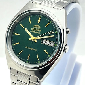 Puede incluir: Un reloj de pulsera Orient Automatic con esfera redonda de color verde oscuro, marcadores de hora y manecillas doradas. El reloj tiene una caja y una correa de eslabones de acero inoxidable, con ventana de día y fecha en WED 13.