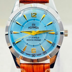 Op de afbeelding: Een Roamer polshorloge met een lichtblauwe wijzerplaat, goudkleurige wijzers en goudkleurige uurmarkeringen. De wijzerplaat bevat de tekst RW ROAMER, 17 JEWELS, SHOCK PROOF en SWISS MADE. Het horloge heeft een zilverkleurige metalen kast en een oranje lederen band.