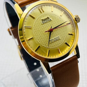 Könnte beinhalten: Eine goldfarbene Armbanduhr mit braunem Lederarmband. Das Zifferblatt zeigt den Text "hmt SONA" und "PARA SHOCK 17 JEWELS". Die Uhr hat ein strukturiertes Zifferblatt und goldene Zeiger mit einem roten Sekundenzeiger.