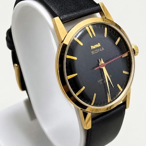 May include: A vintage HMT Sona watch with a black face, gold hands and markers, and a red second hand. The watch has a gold bezel and a black leather strap. The watch is displayed on a white stand.