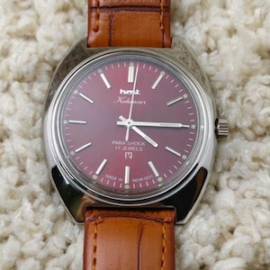 May include: A vintage wristwatch with a burgundy face and silver casing. The watch has a brown leather strap and features the text "hmt Kohinoor" and "Para Shock 17 Jewels". The watch is marked "Made in India".