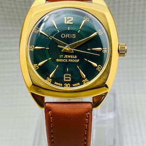 Könnte beinhalten: Eine goldfarbene Armbanduhr mit grünem Zifferblatt und braunem Lederarmband. Das Zifferblatt zeigt die Marke "ORIS", den Text "17 JEWELS SHOCK PROOF" und die Zahlen 12, 6, 30, 45 und 15. Die Uhr hat goldene Zeiger und eine goldene Krone.