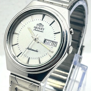 Puede incluir: Un reloj de pulsera automático Orient de tono plateado con esfera redonda blanca, marcadores de hora plateados, manecillas luminosas y una ventana de día y fecha que muestra SAT 10. Incluye una correa de eslabones de acero inoxidable.