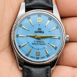 May include: A silver Roamer watch with a light blue face and gold hands. The watch has a black leather strap and the text "17 JEWELS SHOCK PROOF". The watch face displays the numbers 12, 6, 30, and 25. The watch is Swiss made.