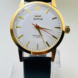 Könnte beinhalten: Eine goldfarbene Armbanduhr mit weißem Zifferblatt und schwarzem Lederarmband. Das Zifferblatt der Uhr hat goldfarbene Markierungen, den Markennamen "SONA" und den Text "PARA SHOCK 17 JEWEL". Die Uhr hat einen roten Sekundenzeiger.