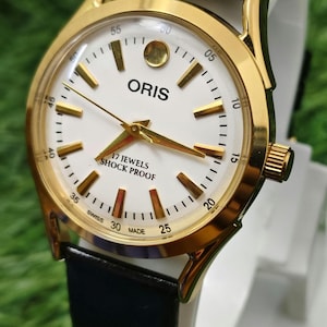Puede incluir: Un reloj de pulsera ORIS dorado con esfera blanca y correa de cuero negro. La esfera del reloj muestra el nombre de la marca, marcadores de tiempo y el texto "17 JEWELS SHOCK PROOF". El reloj tiene agujas doradas y un bisel dorado.
