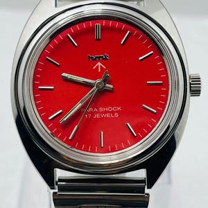 May include: A vintage wristwatch with a bright red face and silver-tone metal band. The watch face features white markers and hands, with the text "PARA SHOCK 17 JEWELS". The watch has a classic, retro design.