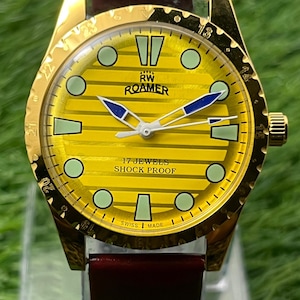May include: A vintage gold-coloured Roamer watch with a yellow dial and a burgundy leather strap. The watch face features green and blue accents, with the text "17 JEWELS SHOCK PROOF" and "SWISS MADE".