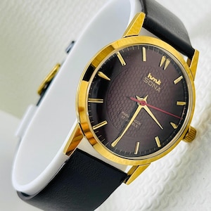 Könnte beinhalten: Eine goldfarbene Armbanduhr mit einem dunkelbraunen Zifferblatt und einem schwarzen Lederarmband. Das Zifferblatt der Uhr hat goldene Stundenmarkierungen und Zeiger, mit dem Text "hmt SONA" und "PARA SHOCK 17 JEWELS". Die Uhr wird auf einer weißen Oberfläche präsentiert.