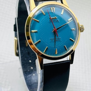 May include: A vintage wristwatch with a gold-coloured case and a textured teal dial. The watch features gold hands and markers, a red second hand, and a black leather strap. The dial reads "SONA" and "PARA SHOCK 17 JEWELS".